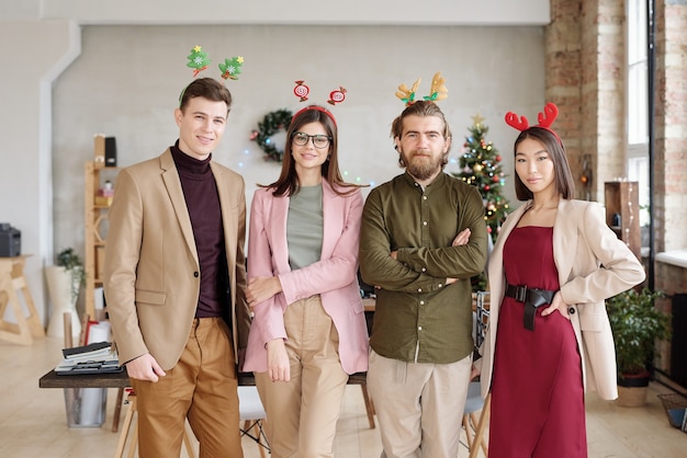 Row of four young intercultural white collar workers in smart casualwear and xmas headbands standing in front of camera in modern office