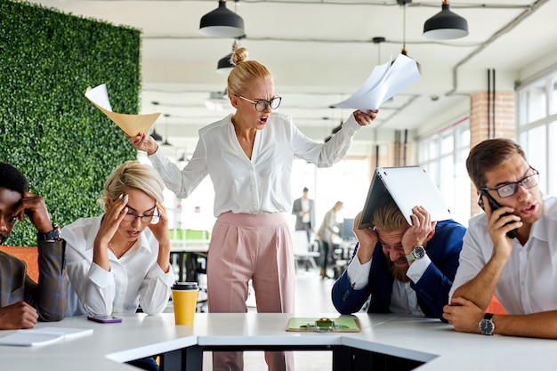 Rude angry female boss executive is dissatisfied irritated by employees, scream at them, incompetent workers. in modern office