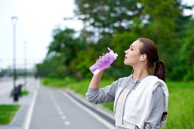 Running asian woman is having break, drinking water during run in summer park