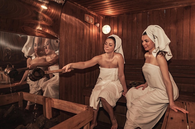 Russian bathhouse Females with bath besoms resting on bench in spa complex Two women relaxing and sweating in wooden sauna