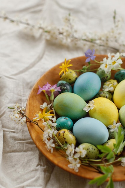 Rustic easter still life Stylish easter eggs and blooming spring flowers in wooden bowl on linen fabric Happy Easter Natural painted eggs and blossoms on rural table