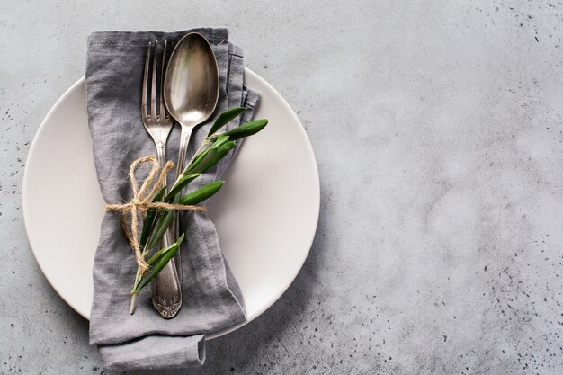 Rustic vintage set of cutlery. Plate with grey linen napkin, fork and spoon, olive tree branch over rustic concrete gray old. Fall holiday table decoration setting. 