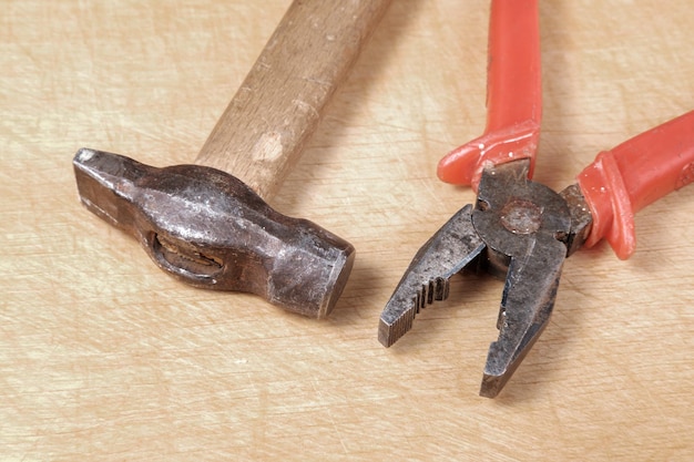 Rusty pliers with a red winding and a hammer with a metal tip and a wooden handle lie parallel to each other on top of the board