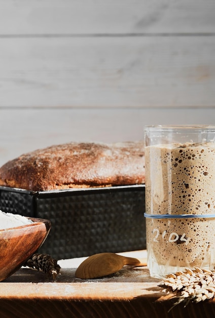 Rye and wheat sourdough starter in a glass jar next to the ingredient flour and freshly baked whole grain bread Close up selective focus in the glass vertical frame