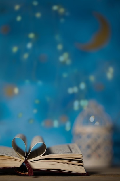 Sacred book of Quran on a wooden surface Blue night sky with crescent moon and candle holder in the background