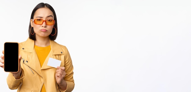 Sad asian girl in sunglasses showing smartphone app interface credit card looking disappointed standing over white background