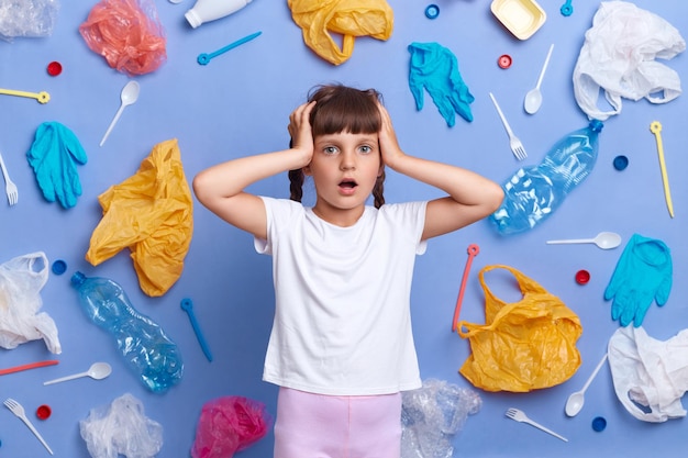 Sad astonished little girl wearing white t shirt standing with hands on head being shocked of pollution on Earth posing against blue wall and much plastic garbage around