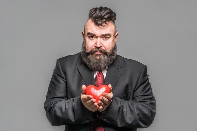 Photo sad bearded man in a black jacket and red tie holding a red heart gray background