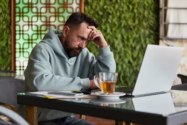 Sad bearded man in casual clothes in cafe using his laptop