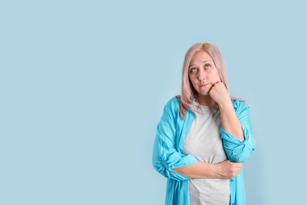 Sad blonde woman holds her fist to her chin and looks up wearily standing on a blue background, copyspace