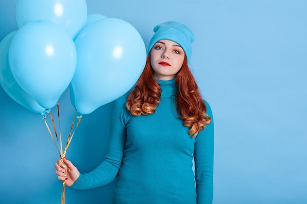 Sad and bored young European woman holding bunch of helium balloons while standing against blue space