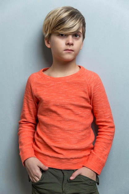 A sad boy of 910 years old stands near a gray wall The guy in the orange sweater keeps his hands in his pockets Loneliness and depression Vertical