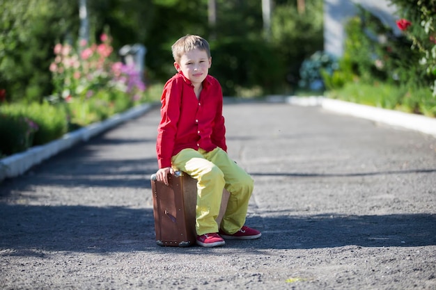 A sad boy sits on an old suitcase on the road The child is about to leave