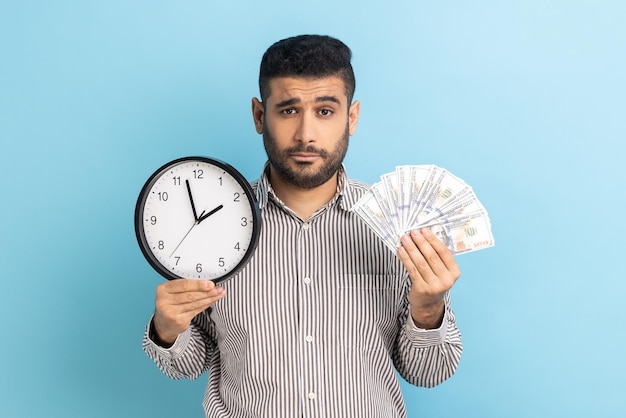 Sad businessman holding big fan of dollar banknotes and wall clock time is money looking at camera