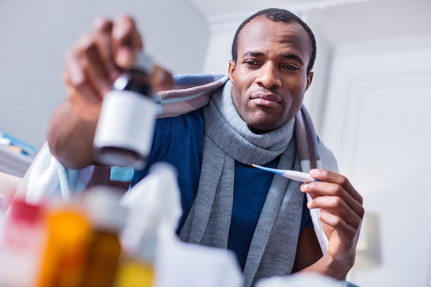 Sad cheerless adult man looking at the bottle with medicine and thinking about illness while holding a thermometer