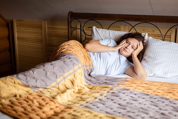 Sad curly brunette woman lying on bed under terry covers with worried face having inner problems wearing home pajamas in daytime Anxiety moment in adult life