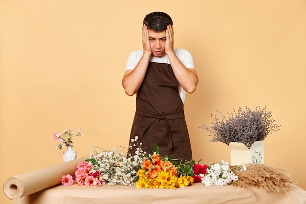 Sad despair man florist making bouquet in flower shop standing isolated over beige background having lots order has not enough time