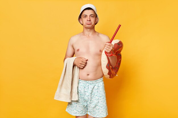 Sad disappointed man wearing shorts swimsuit and panama holding donut rubber ring isolated on yellow background looking at camera with pout lips expressing sorrow while resting near beach