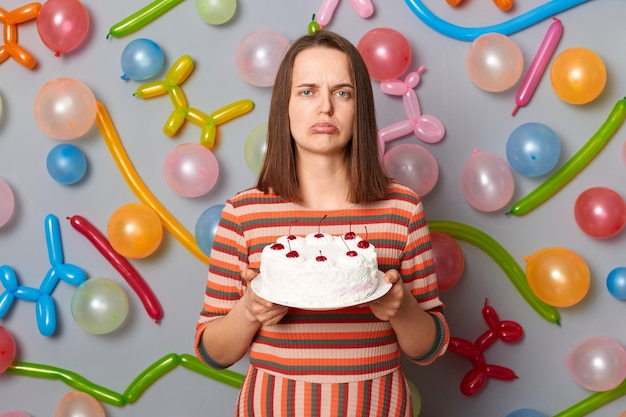 Sad disappointed woman with brown hair wearing striped dress holding cake she dislikes mixing order buying wrong dessert standing against gray wall decorated with colorful balloons