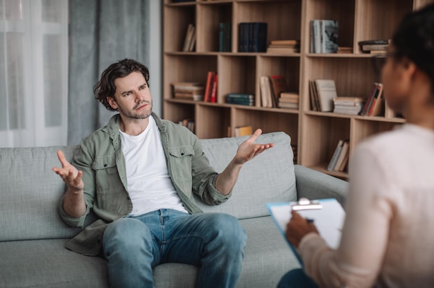 Sad dissatisfied european man spreads his arms to the sides and listens to black female psychologist