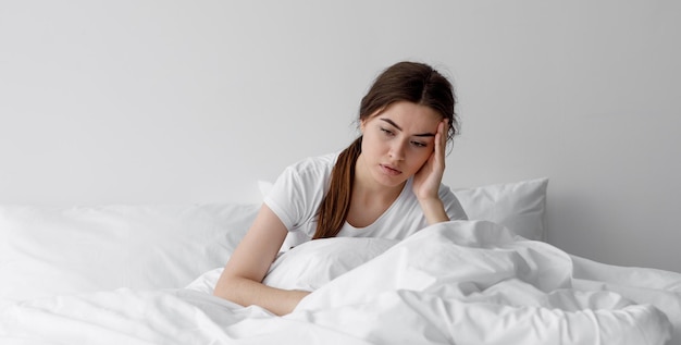 Sad european millennial female worried suffers from insomnia and headache Lady sits on bed in bedroom