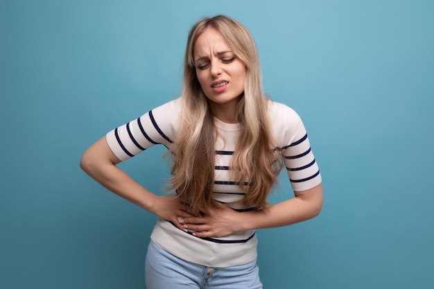 Sad girl in casual clothes clutches her stomach because of indigestion on a blue isolated background