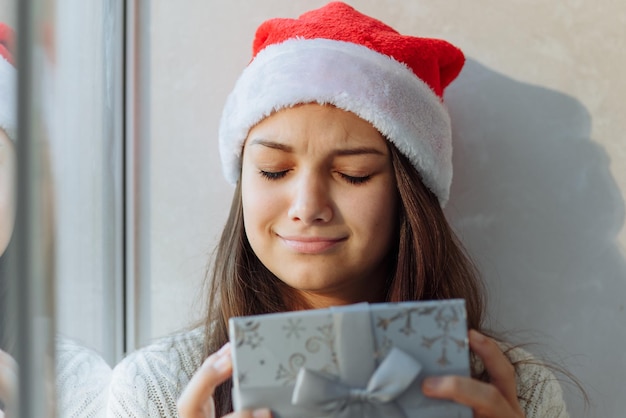 sad girl in a Christmas hat with a gift box by the window closeup