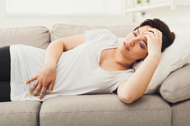 Sad girl with headache or stomachache. Young brunette woman feeling pain, lying on sofa at home