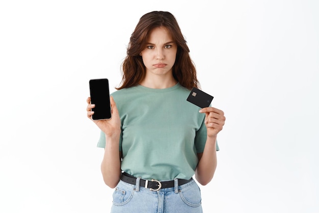 Sad and gloomy young girl showing smartphone empty screen and plastic credit card empty bank account balance on mobile banking app standing against white background