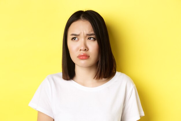 Sad and jealous asian girl sulking, frowning and looking left with upset face, standing over yellow background.