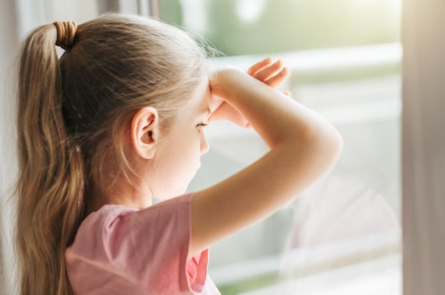 Sad little girl looking through window at home