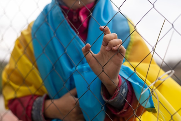 Sad little girl with the flag of Ukraine behind a metal fence. Social problem of refugees and forced migrants