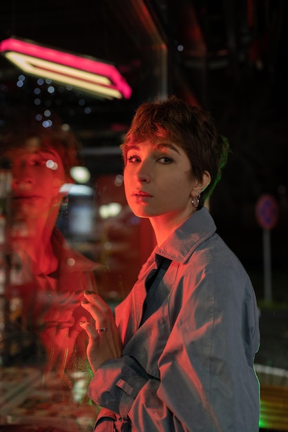 Sad lonely woman in red neon light leaning on shop window on street at night looking at camera