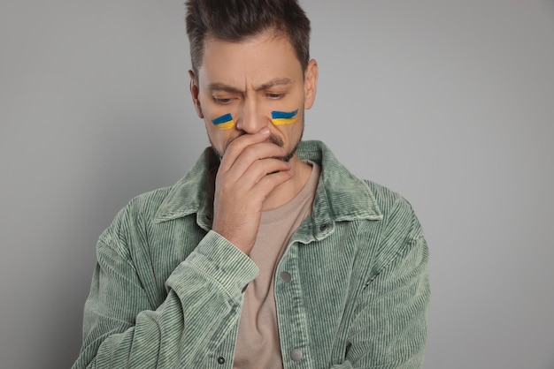 Sad man with drawings of Ukrainian flag on face against light grey background