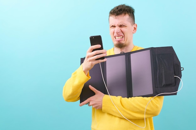 A sad man with a grimace holds a foldable portable solar panel and looks at the phone on blue