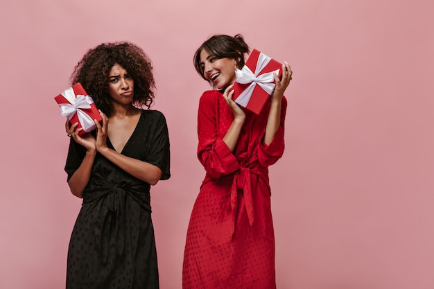 Sad mulatto woman in dark outfit looking into camera, holding red gift box and posing with happy girl in bright clothes