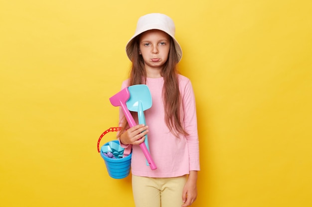 Sad offended little cute girl in panama holding beach sandbox toys rake and shovel isolated over yellow background looking at camera with pout lips felling unhappy and bored