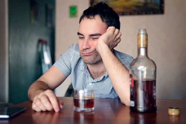 Sad sad young alcoholic sit at the table with a bottle of rum. the face of a drunk man. Stupid emotion. Depression and alcoholism.