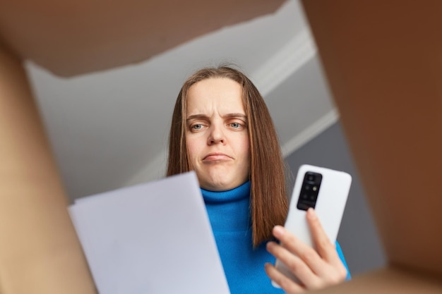 Sad serious woman customer wearing blue turtleneck opening parcel looking at consignment trying to understand where is a mistake holding phone needs to call to delivery service