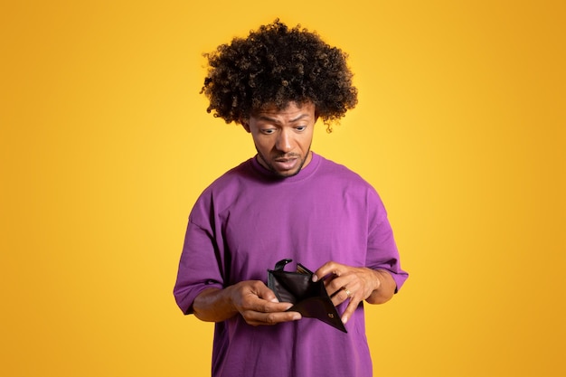 Sad shocked adult african american curly man in purple tshirt looks at empty wallet