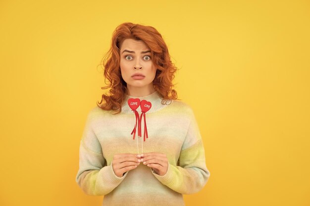 Sad shocked redhead woman with red heart sticks on yellow background valentines day