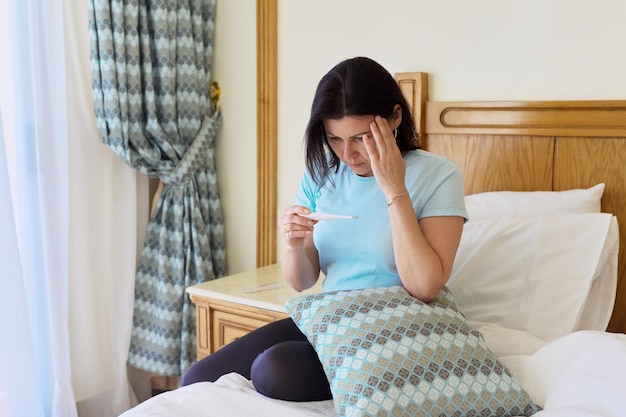 Sad sick mature woman holding thermometer in her hands sitting at home in bed