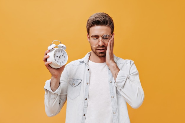 Sad stylish guy in round eyeglasses, white modern shirt and cool t-shirt posing with closed eyes and holding big alarm clock