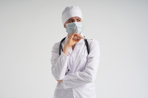 Sad surprised doctor wearing medical face mask holding medical thermometer over white wall