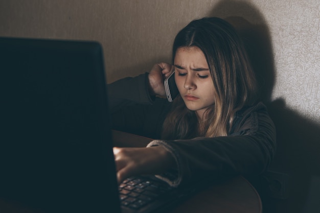 Sad teenage girl sitting near laptop in dark room. She is a victim of online bullying Stalker social networks.