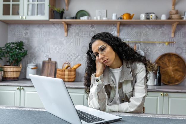 Sad and thinking woman working at home using laptop hispanic woman in kitchen at table wearing