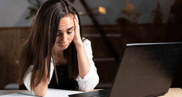 Sad tired Caucasian woman sitting with laptop in office or at home under stress from bad news girl holds her head with her hands unmotivated employee at the workplace Work and health concept