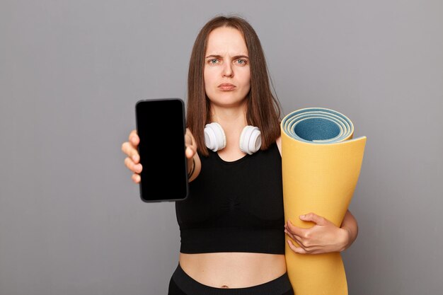 Sad unhappy woman holding rolled fitness mat dressed in activewear isolated over gray background showing mobile phone with empty screen advertisement area