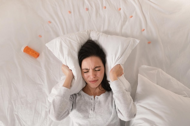 Sad unhappy young european woman lies on white bed with jar and pills covers ears with pillow in bedroom