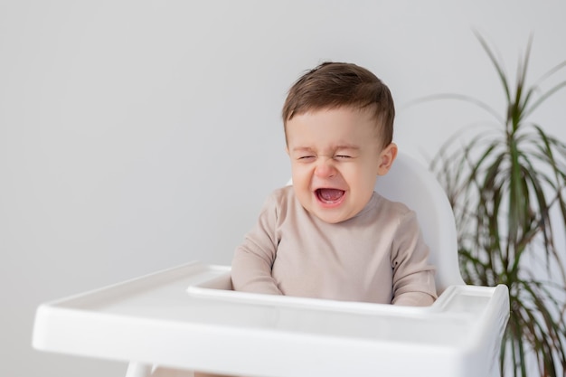 Sad upset baby is sitting in a white high chair for feeding little boy is crying